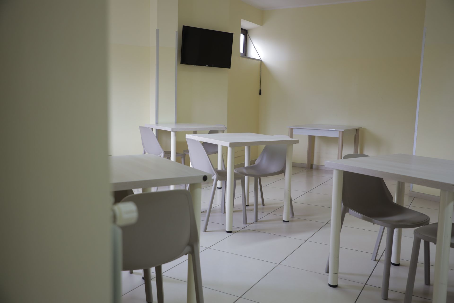 Una stanza con pavimento piastrellato di bianco, diversi tavolini quadrati bianchi e sedie grigio chiaro abbinate, oltre a un televisore a parete.