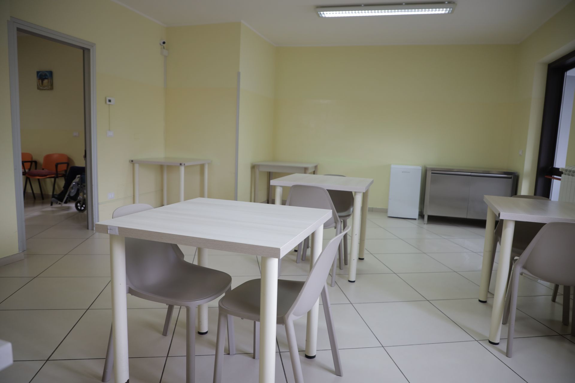 Una stanza luminosa con pareti giallo pallido, pavimento piastrellato e diversi tavoli bianchi con sedie grigie disposti adibiti a zona pranzo.