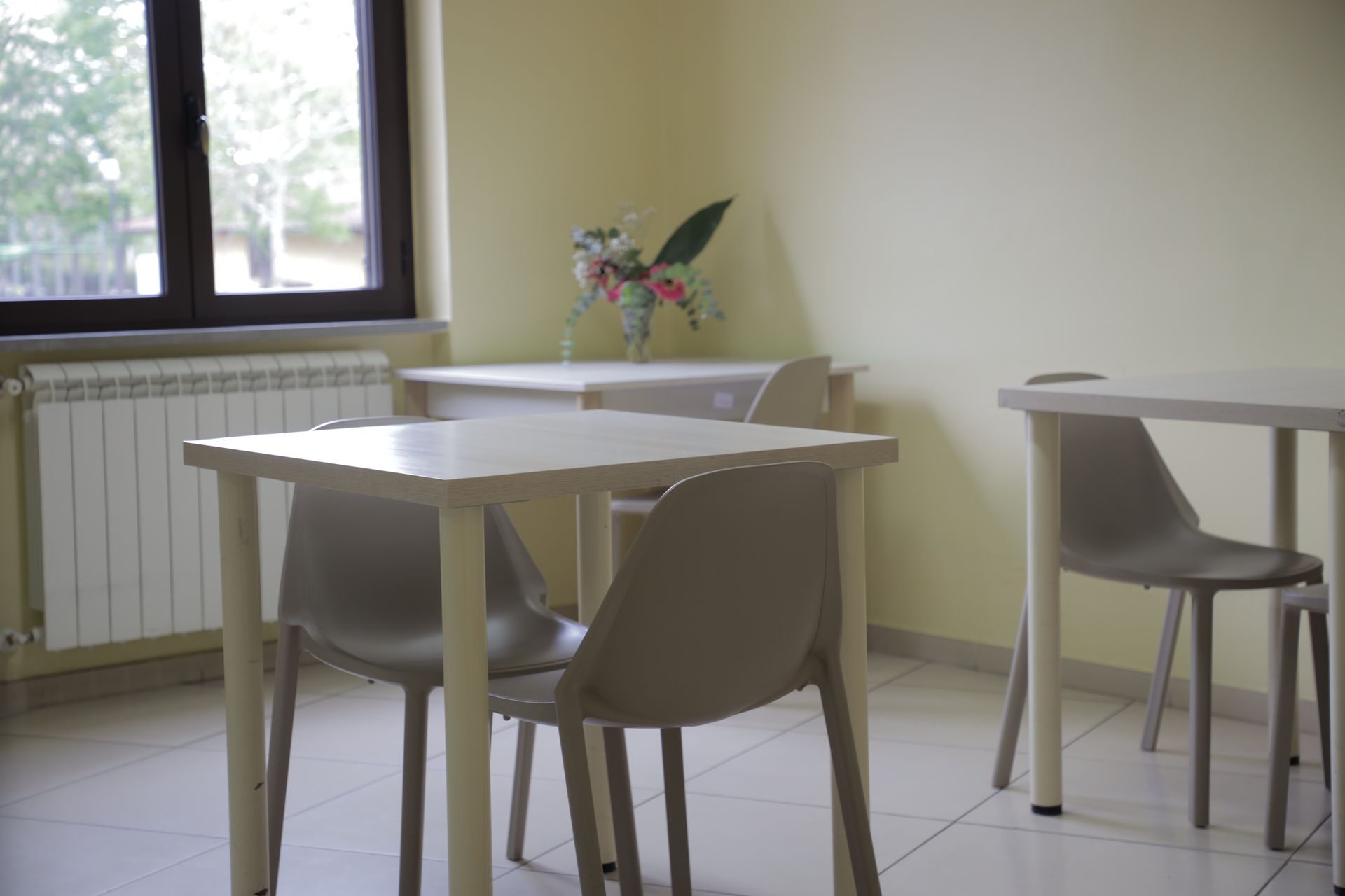 Due tavoli chiari con sedie grigie in una stanza tranquilla con pareti color crema, finestra e termosifone.