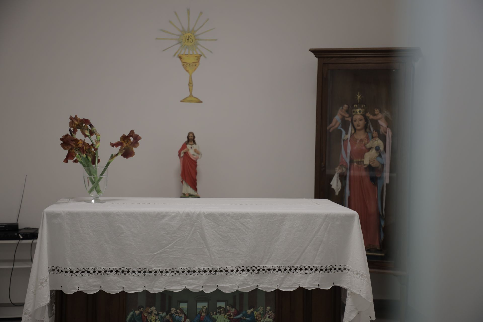 Un altare con una tovaglia bianca bordata di pizzo, un vaso di fiori, una statua di Gesù e un affresco raffigurante un calice e un'ostia.