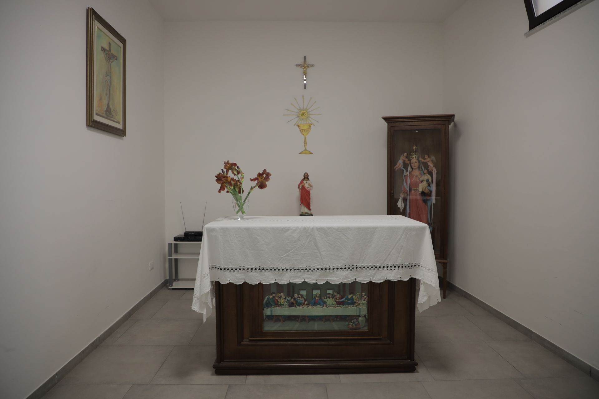 Una piccola cappella con un altare ricoperto da una tovaglia di pizzo bianco, un vaso di fiori, una statua religiosa e delle decorazioni murali.