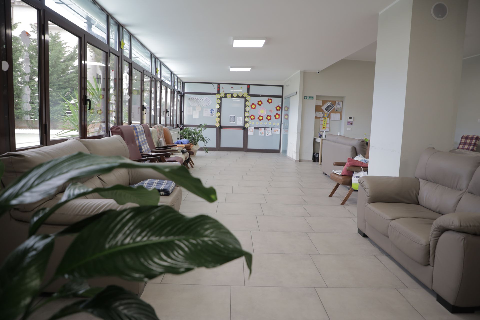 Una sala d'attesa luminosa e moderna con divani chiari, pavimento piastrellato e ampie finestre lungo una parete.