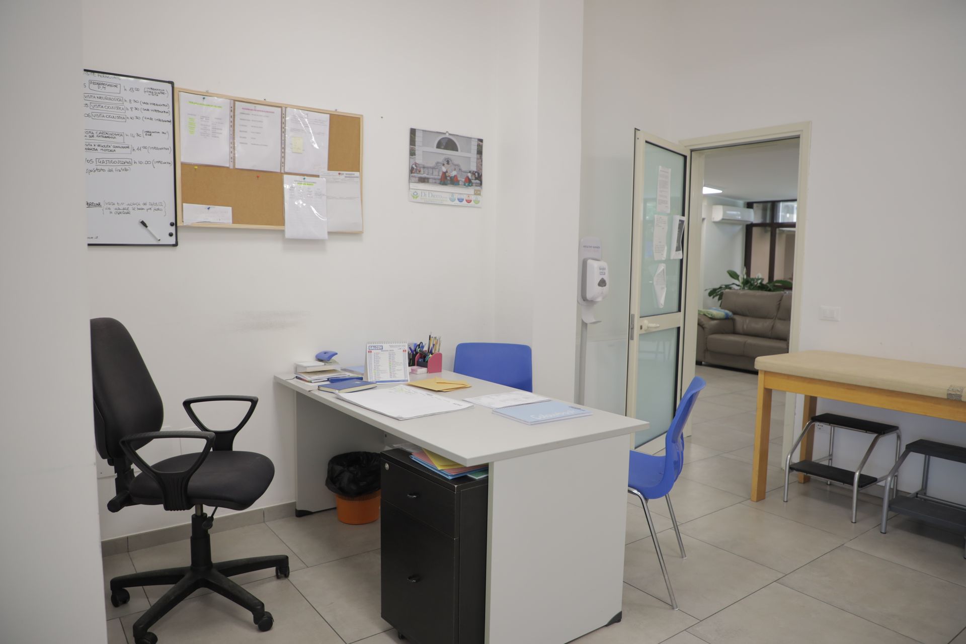 Uno studio medico con scrivania, sedia girevole, sedie blu, bacheca di sughero e lettino per le visite in una stanza separata.