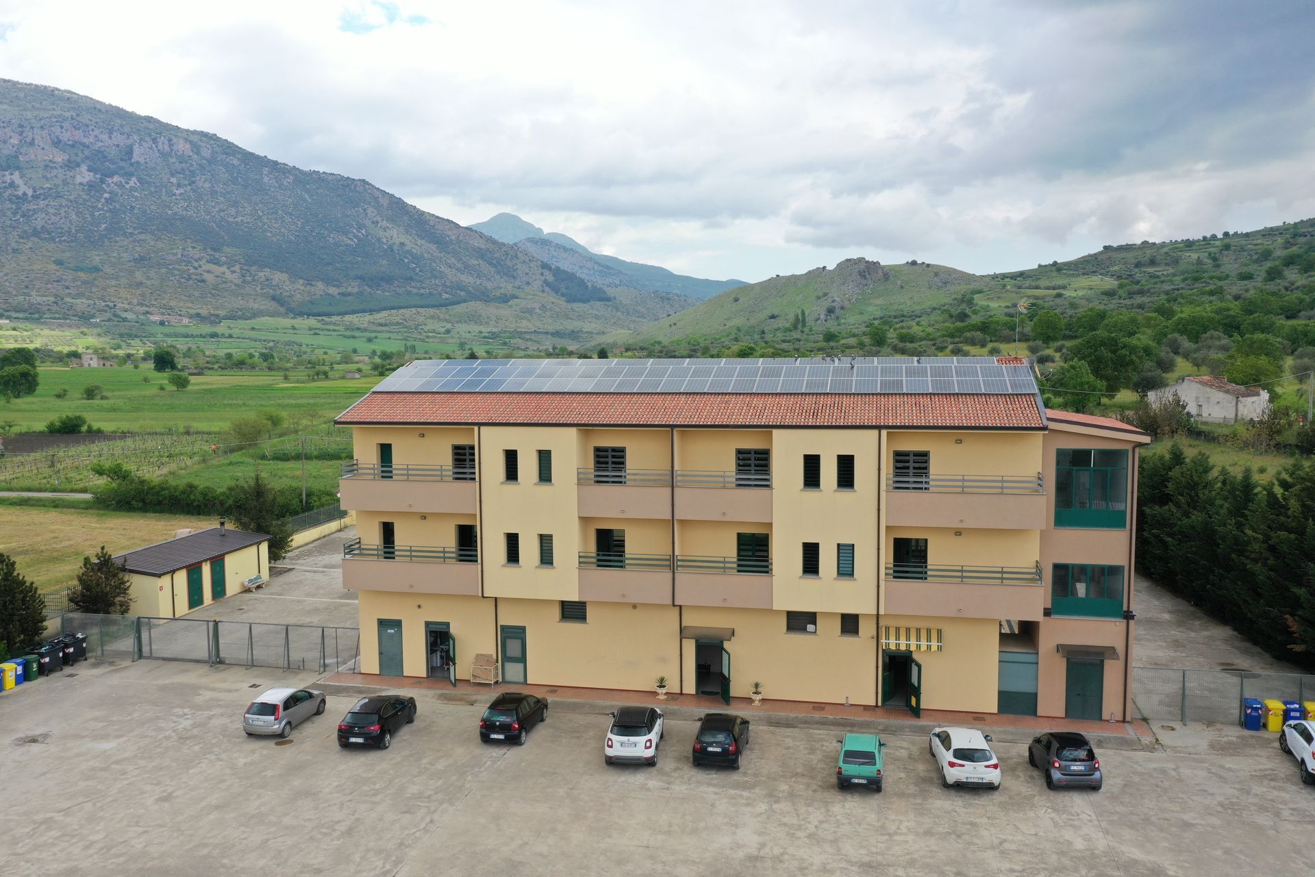 Un edificio beige a tre piani con pannelli solari sul tetto e un parcheggio con diverse auto di fronte alle montagne.
