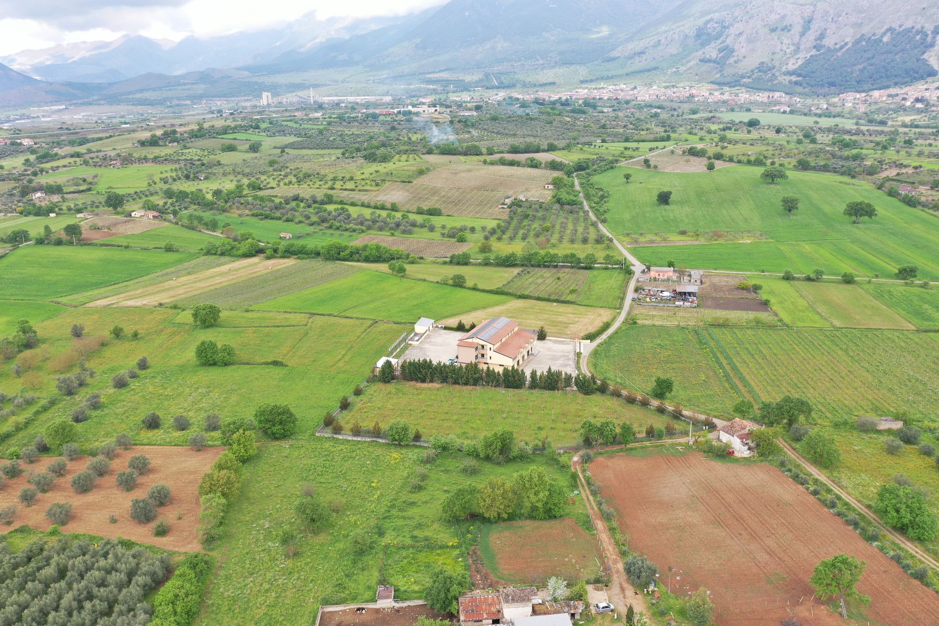 Veduta aerea di un paesaggio rurale con edifici agricoli, campi verdi, frutteti e montagne in lontananza sotto un cielo nuvoloso.