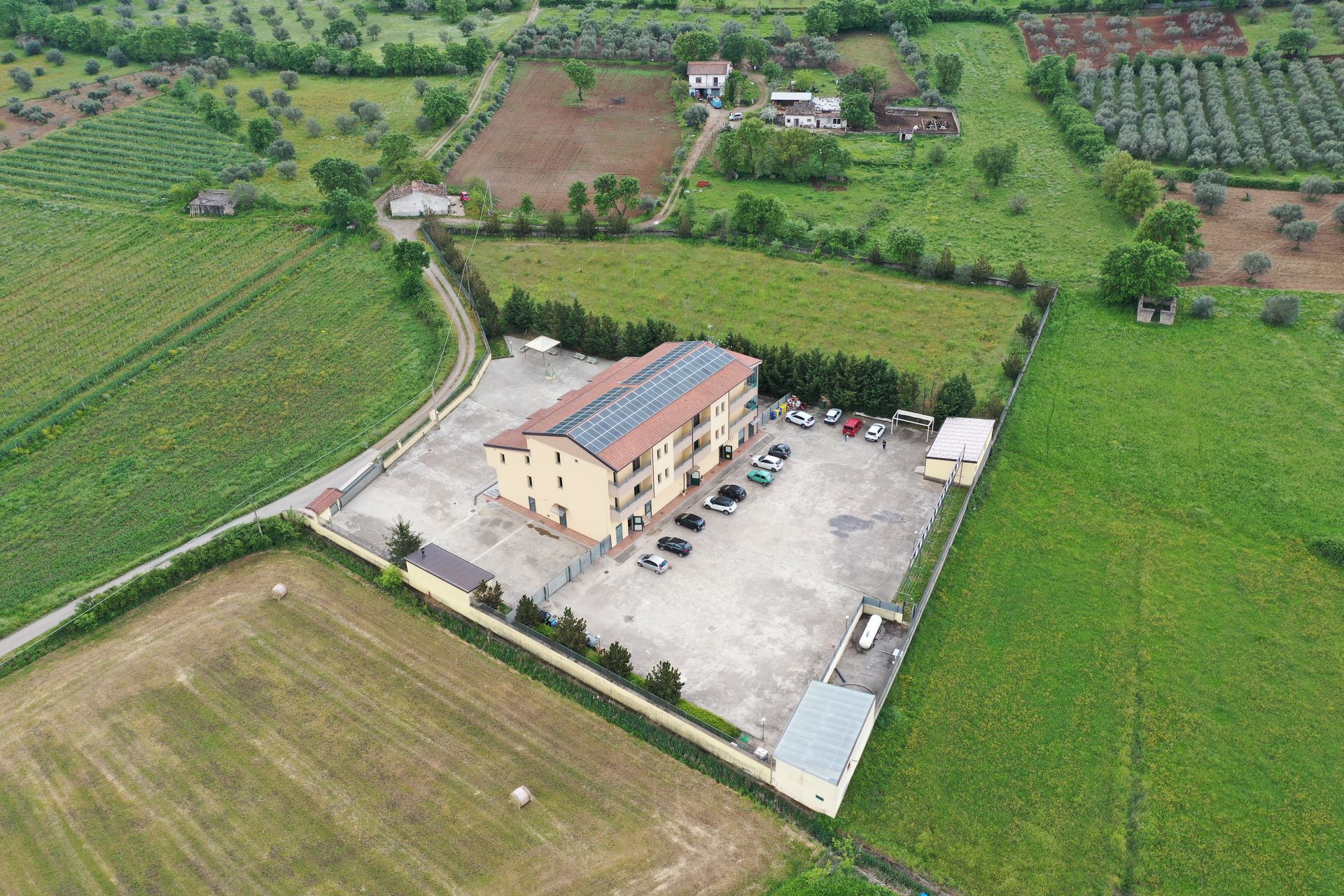 Veduta aerea di un edificio multipiano beige con tetto ricoperto di pannelli solari, situato in un parcheggio circondato da campi verdi.