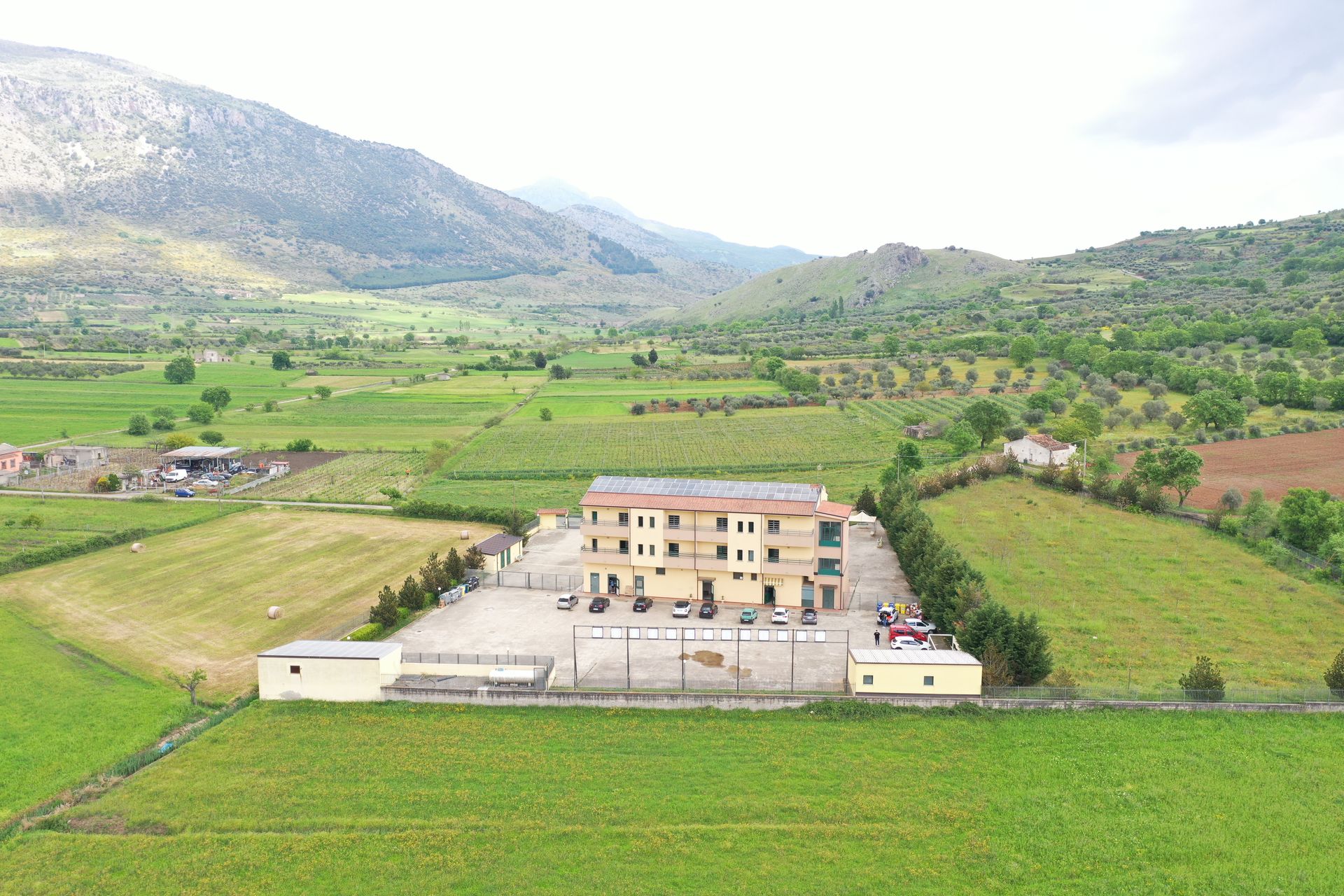 Una vista aerea mostra un edificio beige a tre piani circondato da campi verdi e colline in un paesaggio rurale.