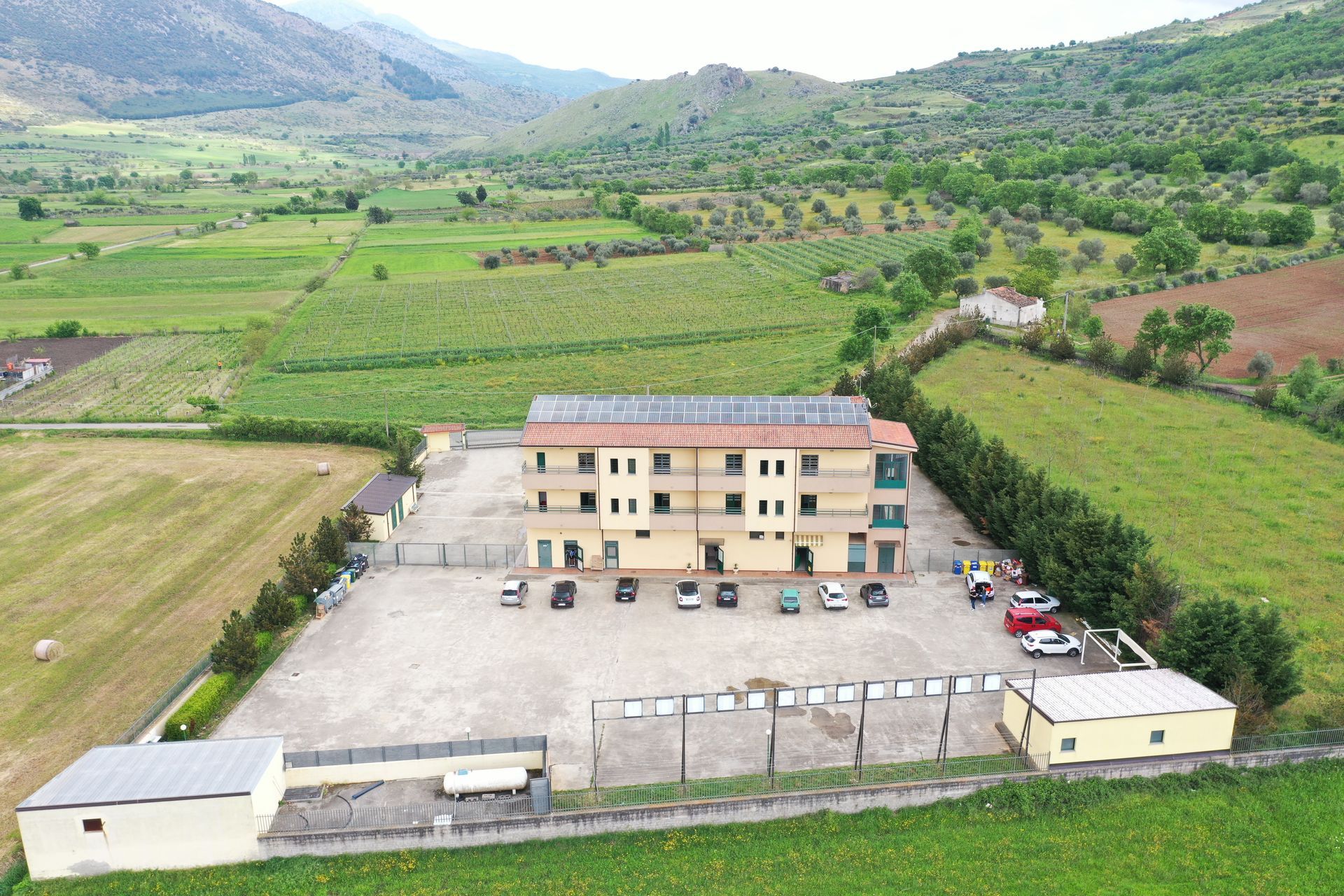 Veduta aerea di un edificio residenziale beige a tre piani, circondato da campi verdi, colline e un parcheggio asfaltato.