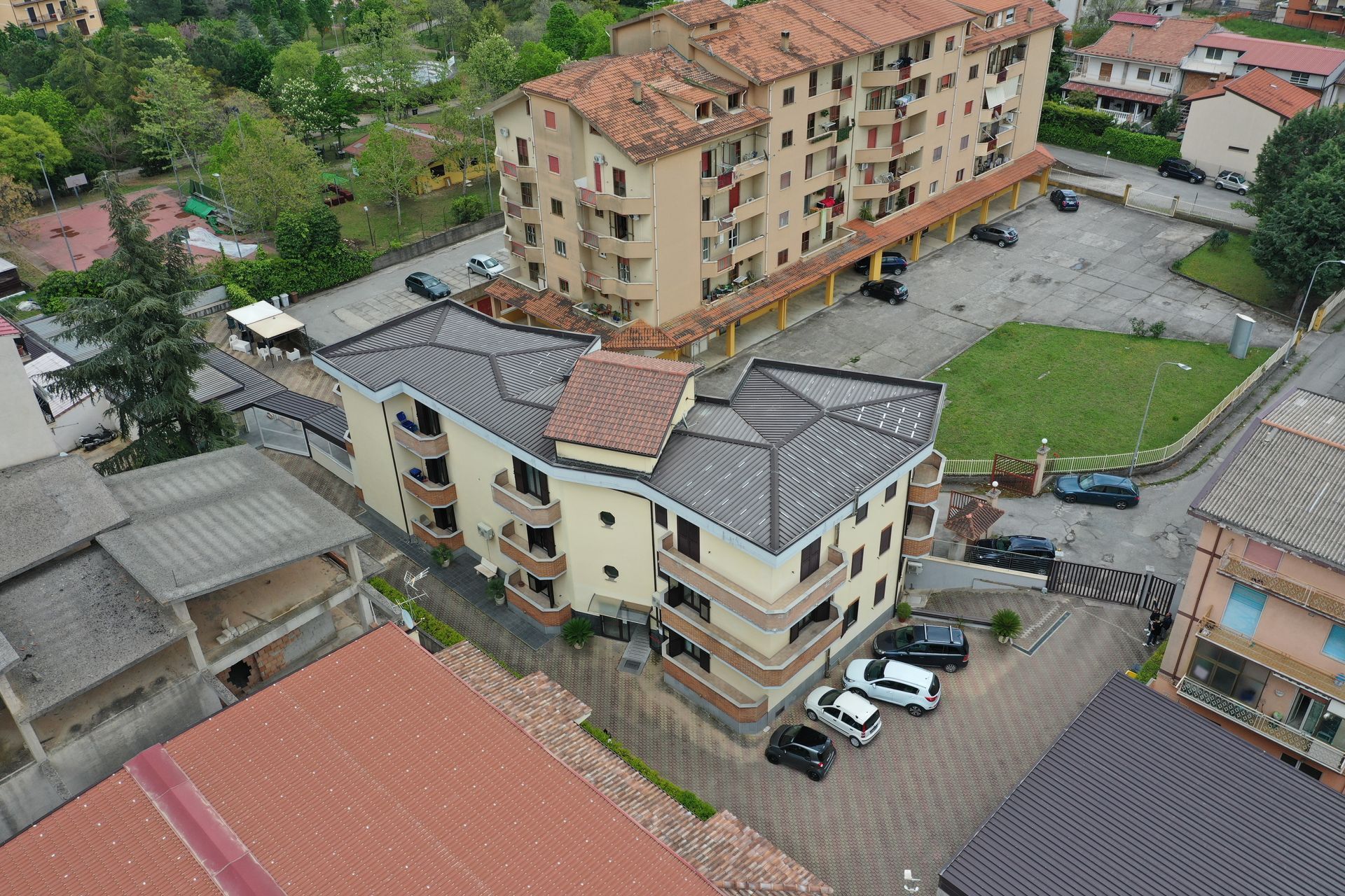 Veduta aerea di un complesso residenziale color crema con tetto di tegole scure, adiacente a un edificio più grande e a un'area parcheggio.