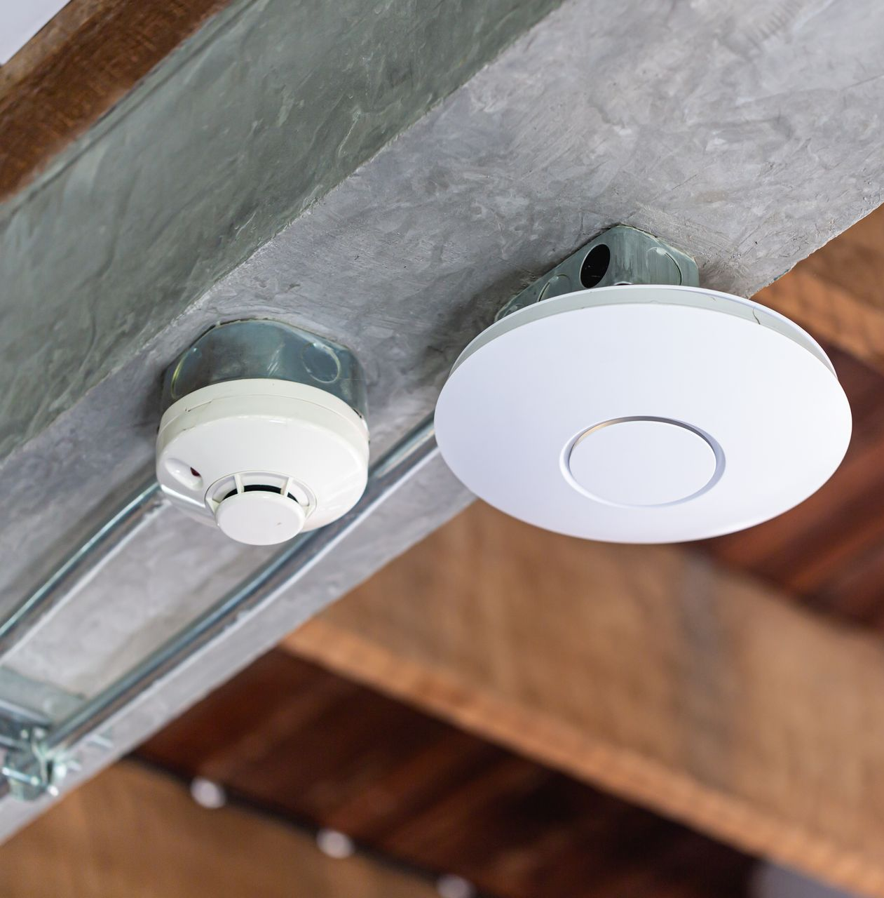 Two white ceiling-mounted smoke detectors on a metal beam under a wooden roof