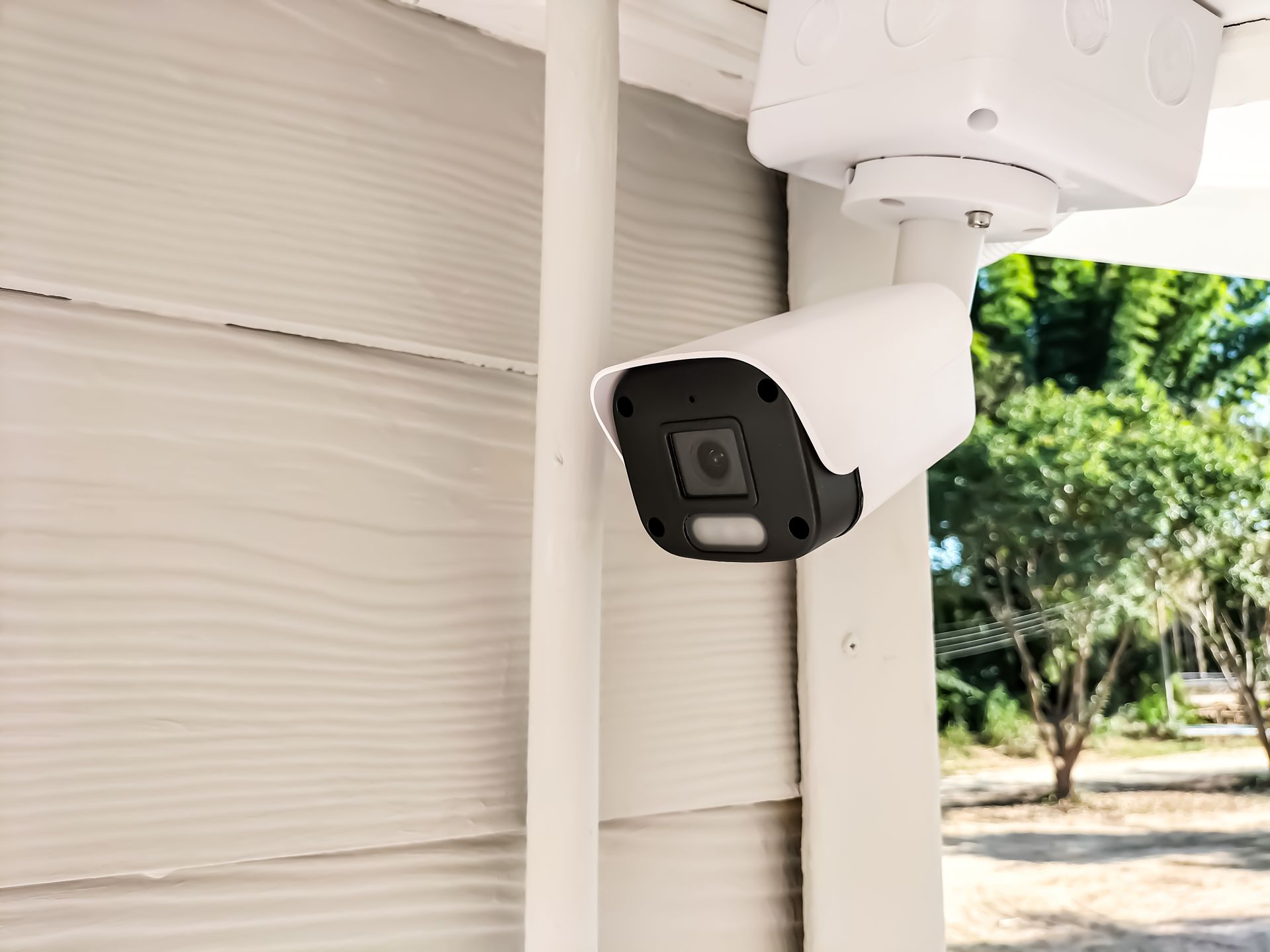 White security camera mounted on a pole beside a house, aimed outdoors near a yard.