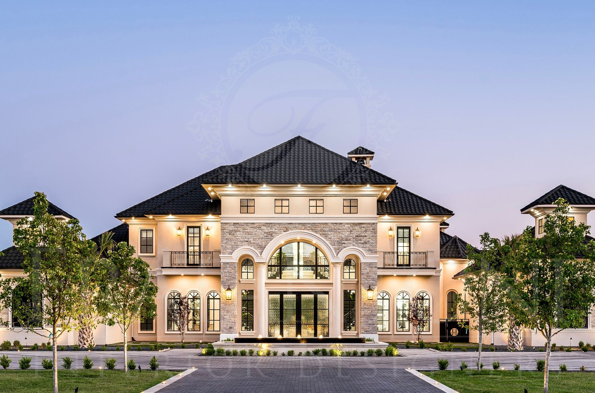 Large, beige and stone mansion at dusk, arched entryway, dark tiled roof, trees line the driveway designed by Fratantoni Interior Designers in Scottsdale Arizona