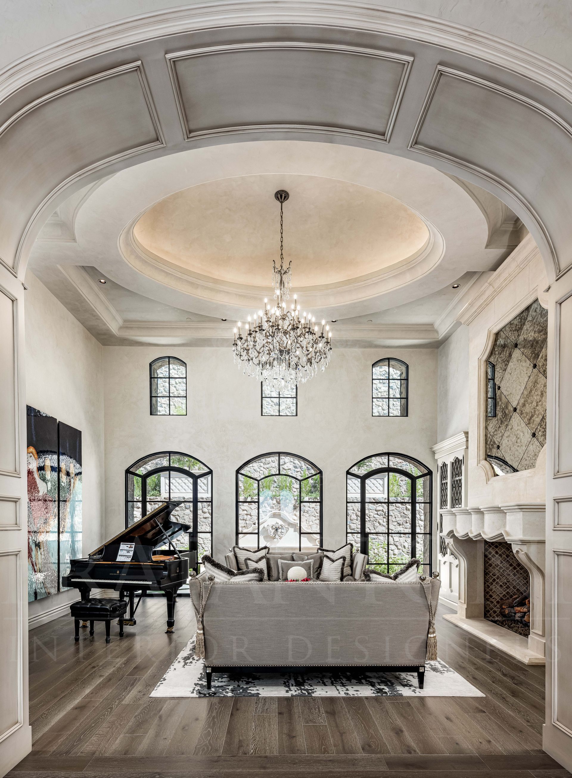 French style luxurious living room interior design with a grand piano, fireplace, arched windows, and a large chandelier under a circular ceiling by Fratantoni Interior Designers