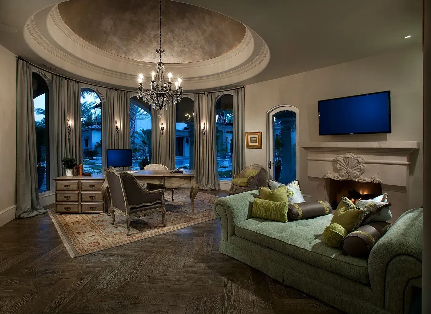 Elegant home office with a luxury chandelier, curved windows, desk, and lounge seating.