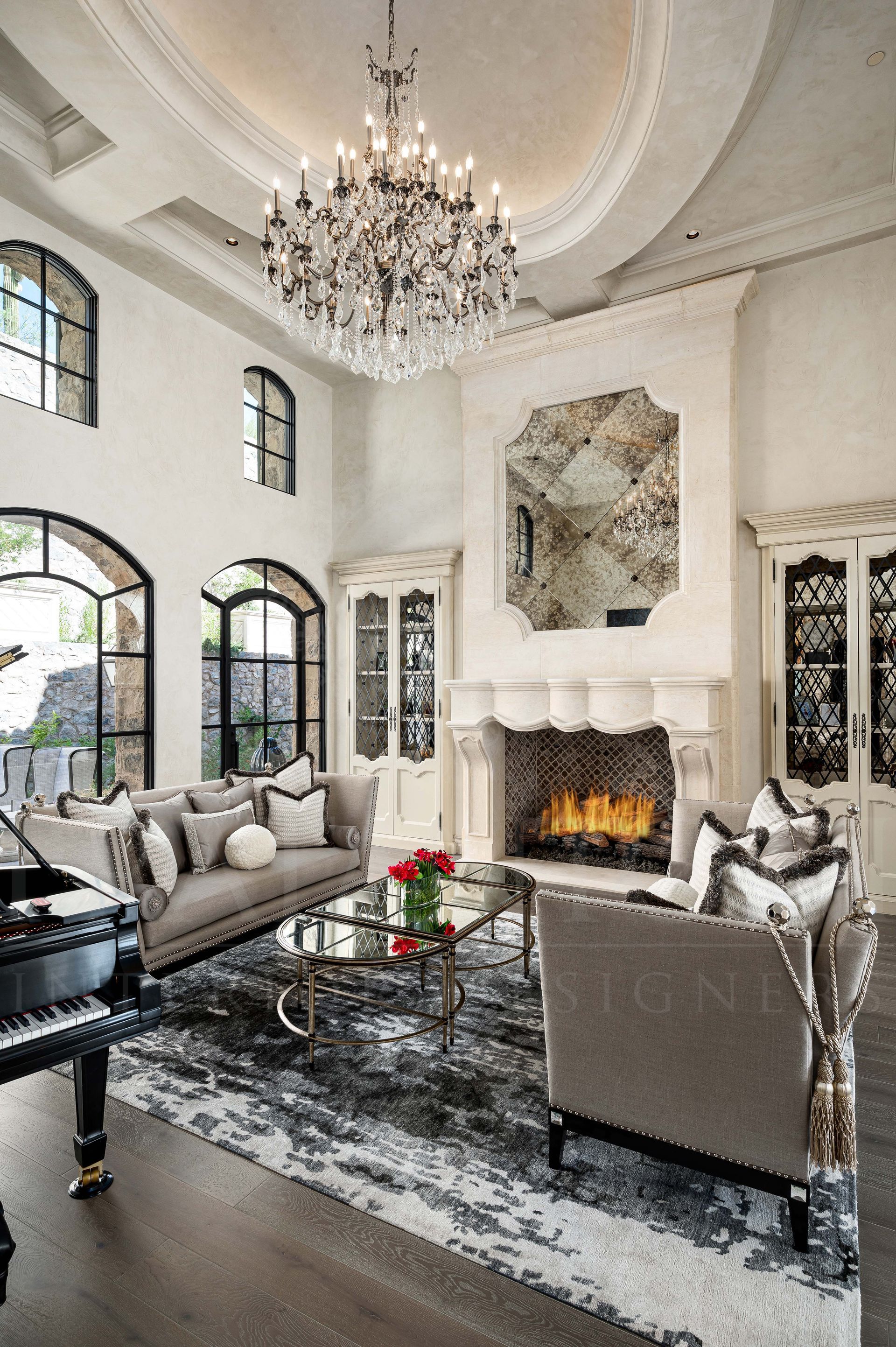 French luxury grand living room featuring a crystal chandelier, carved-stone fireplace, arched windows, and custom grey furniture on a patterned rug.