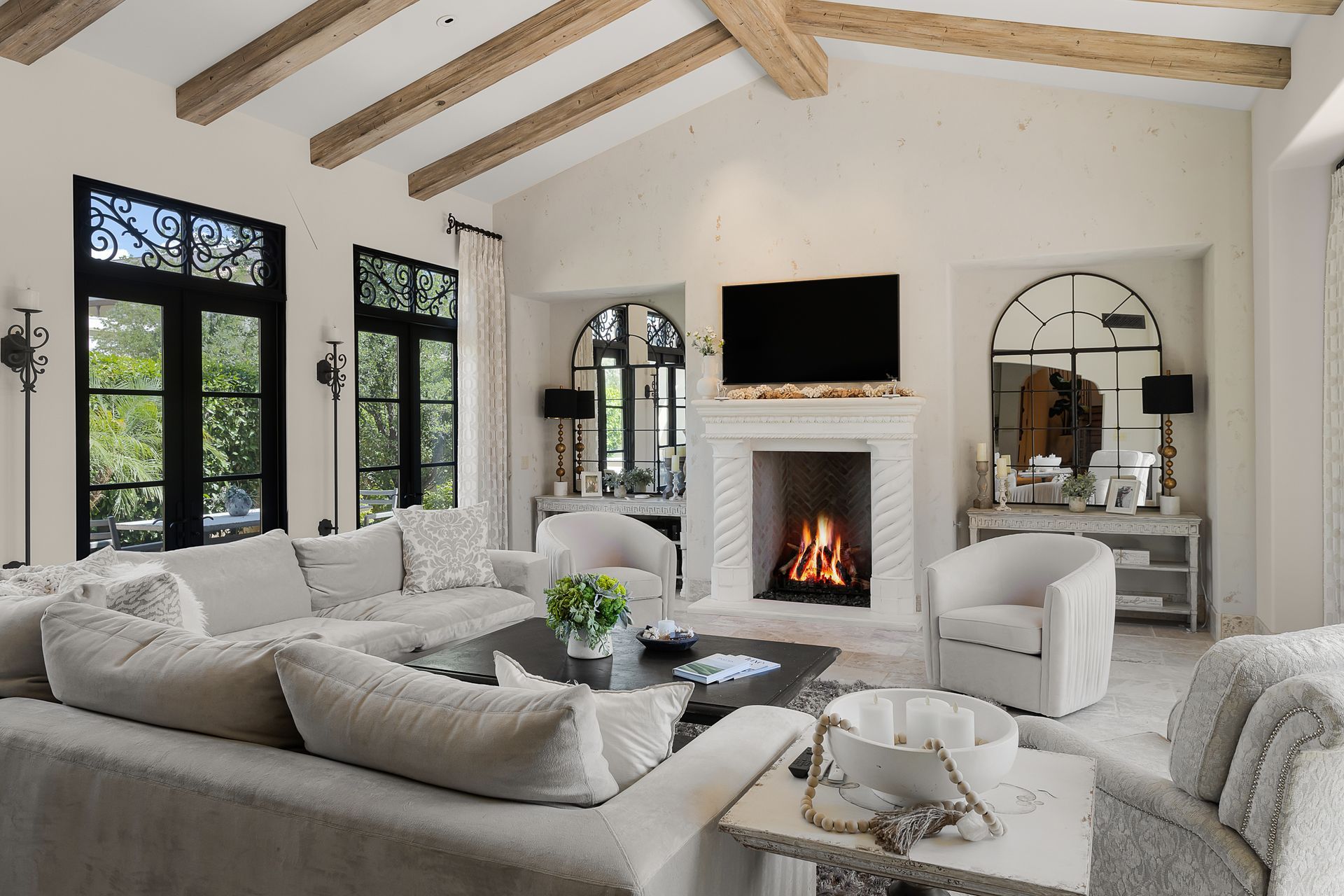 Living room with cream-colored walls, fireplace, and large windows.  Neutral-toned furniture and wooden ceiling beams.