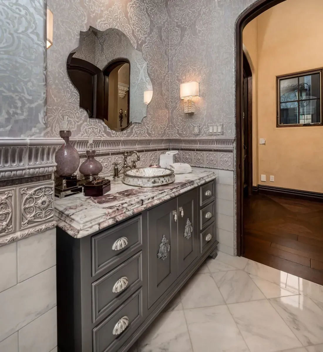 Grey vanity with marble countertop and decorative mirror in a luxurious bathroom with patterned wallpaper.