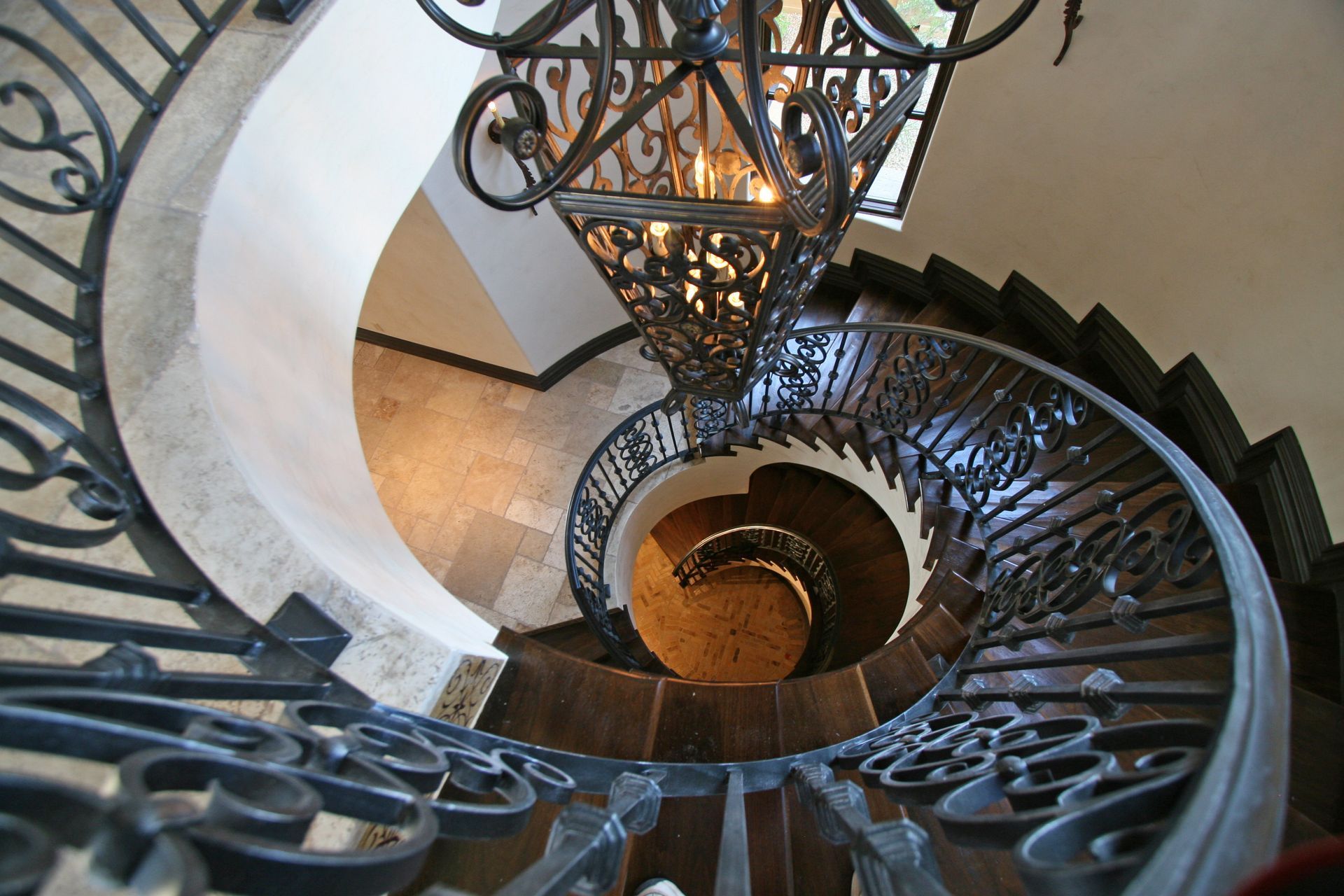 Spiral staircase with ornate wrought iron railing, viewed from above, lit by chandelier.