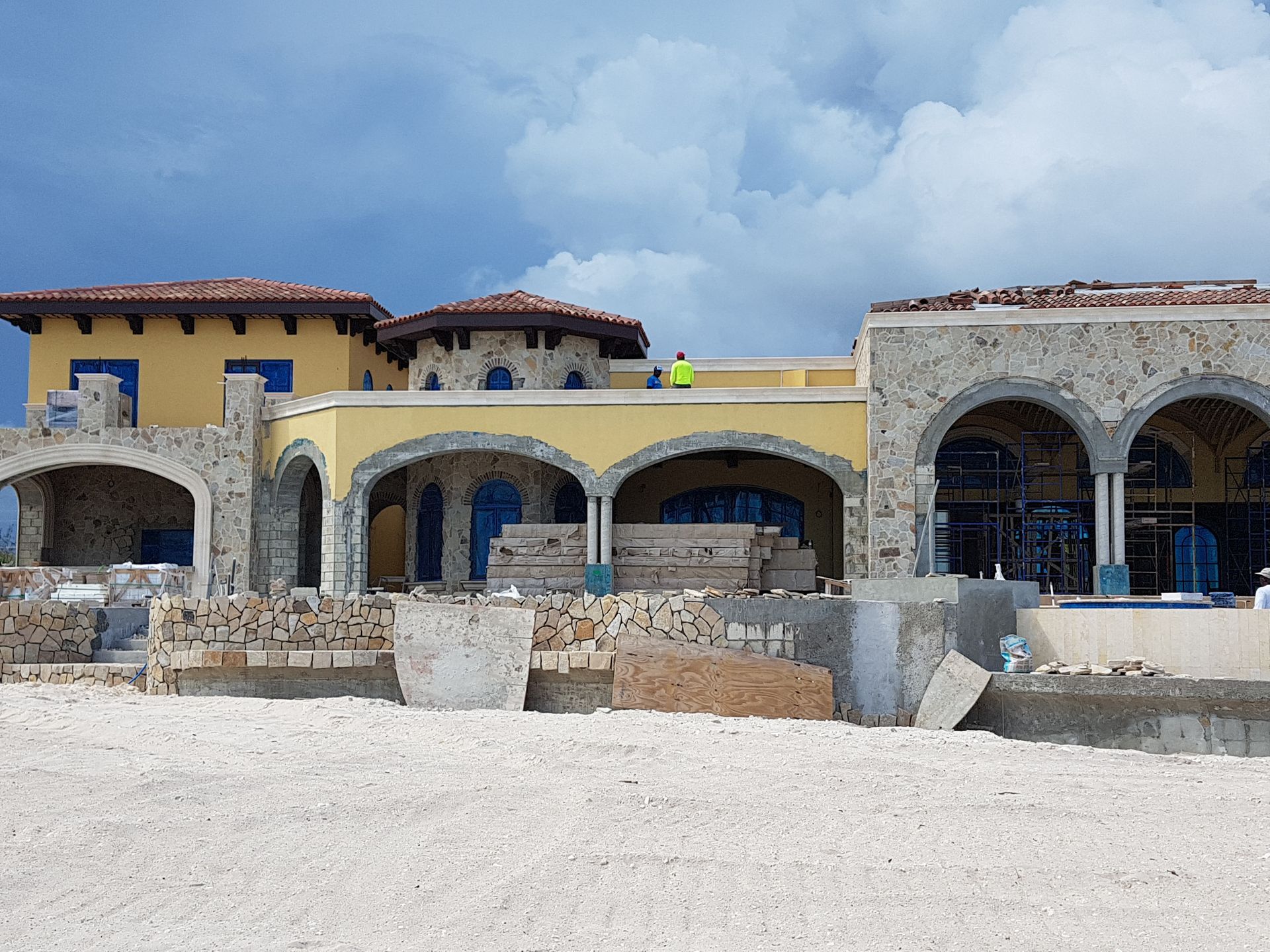 Mediterranean-style building under construction, yellow and stone exterior, arched windows, and a blue sky in the background.