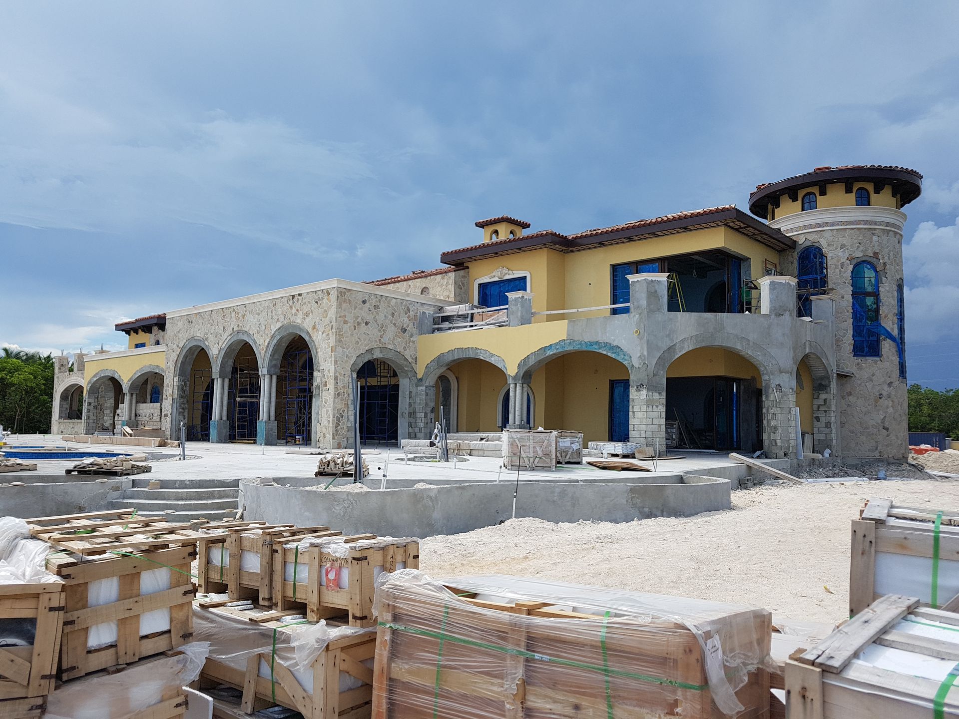 A large Mediterranean-style building under construction, with a tower, arched doorways, and light-colored stone and stucco.