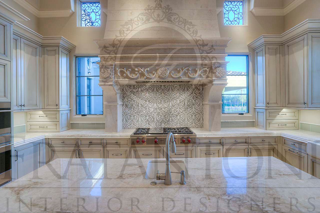 Bright luxury kitchen with marble island, white cabinetry, custom carved stone range hood, and ornate stone backsplash around an island sink.