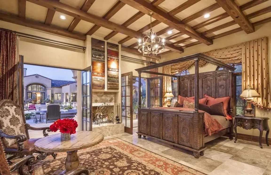 Luxurious bedroom with carved four-poster bed, rug, open doors to patio, chandelier, and a small table.