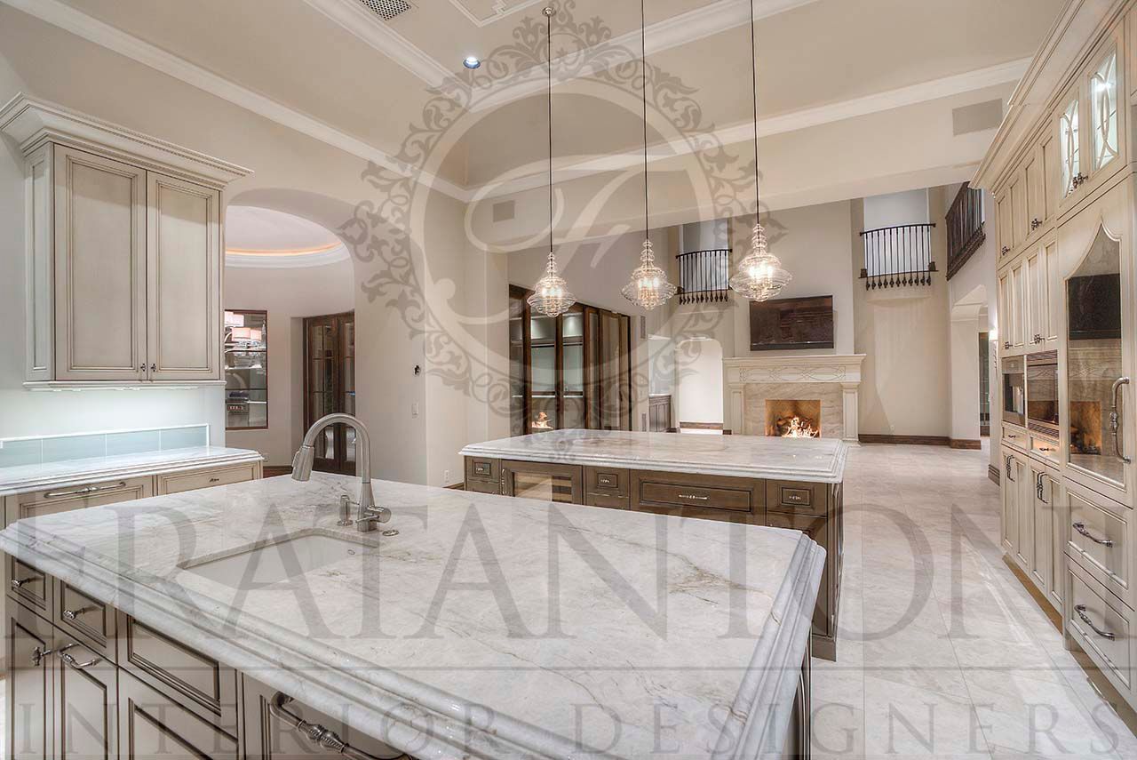 Elegant neutral luxury kitchen with two marble islands, ornate cabinetry, and pendant lights. Custom kitchen attached to great room with fireplace and tall ceilings