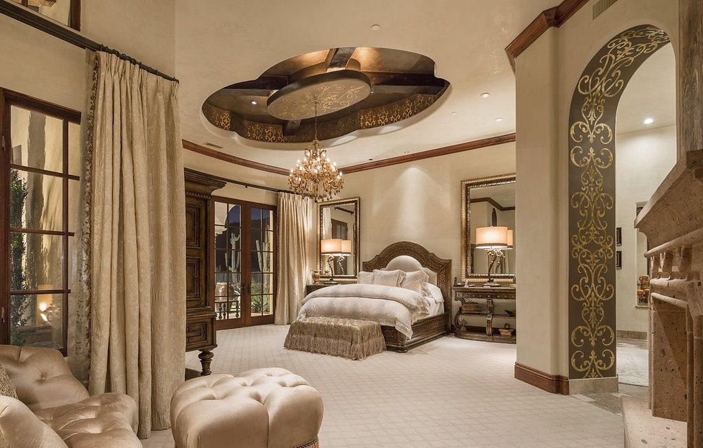 Luxurious bedroom with cream colors, ornate ceiling, chandelier, bed, and decorative archway.