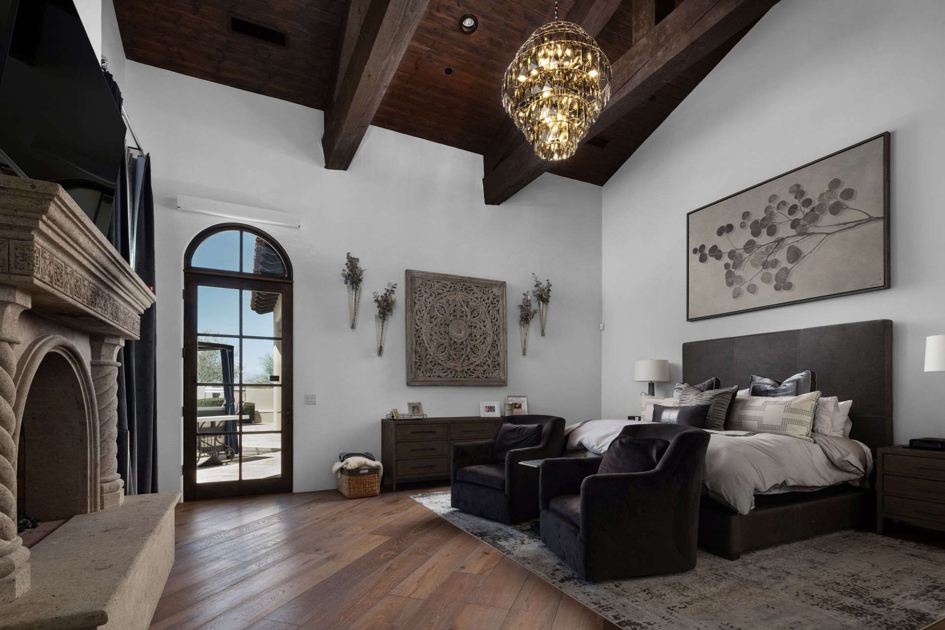 Bedroom with fireplace, wood ceiling beams, decorative chandelier, dark furniture, and outdoor view.