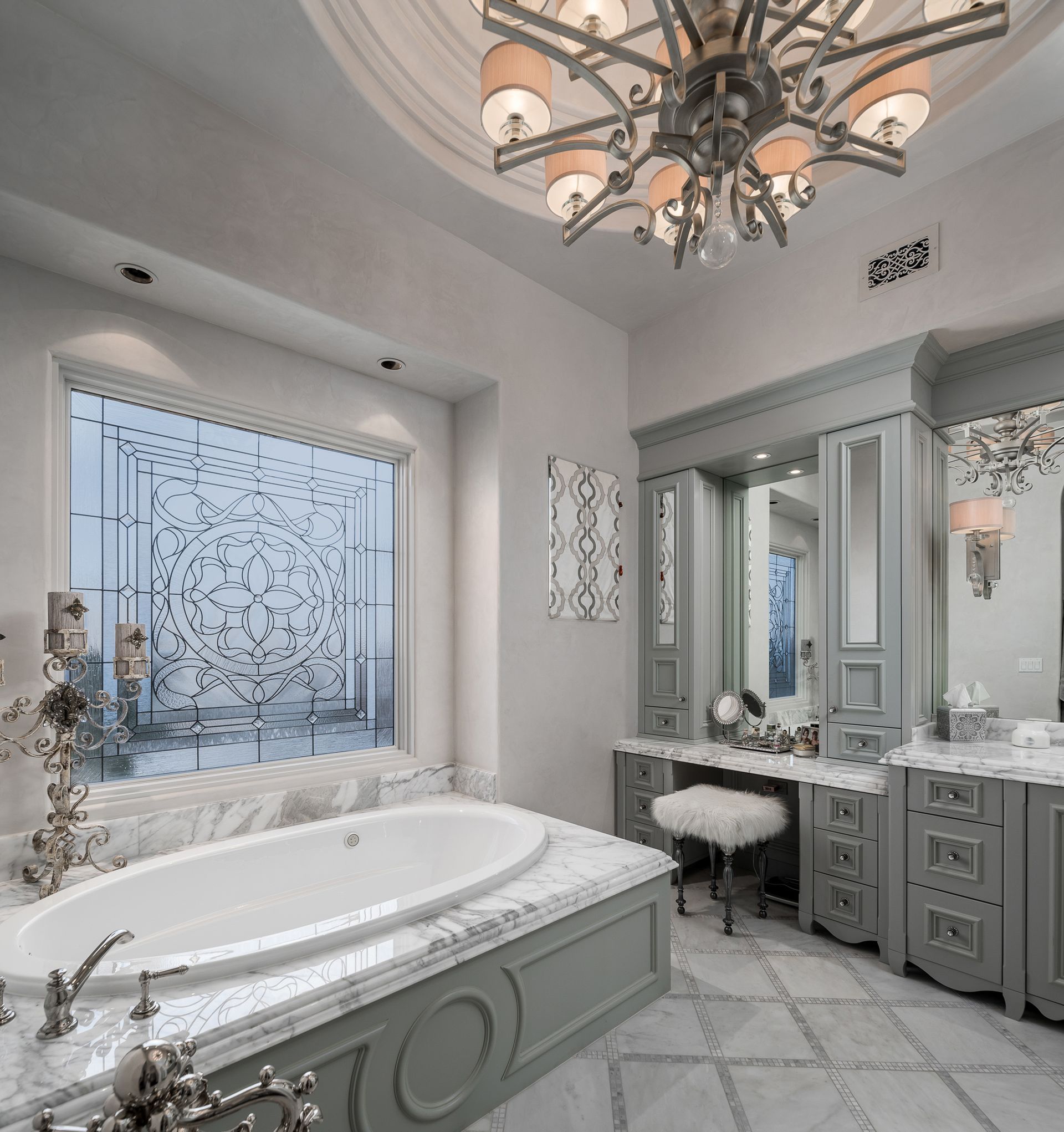 Elegant bathroom with a marble tub, vanity, and chandelier, with a decorative window.