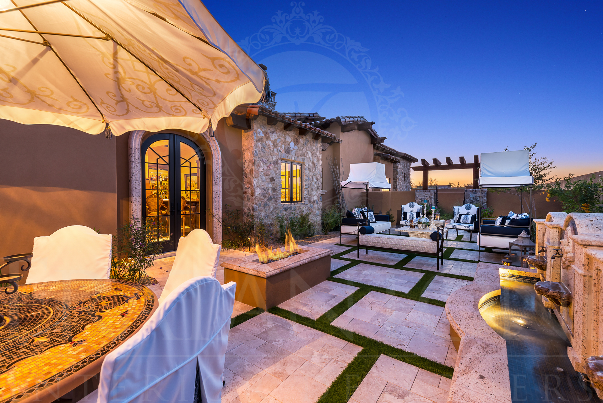 Patio with fire pit, seating, and umbrella at dusk. Stone walls and blue sky backdrop.
