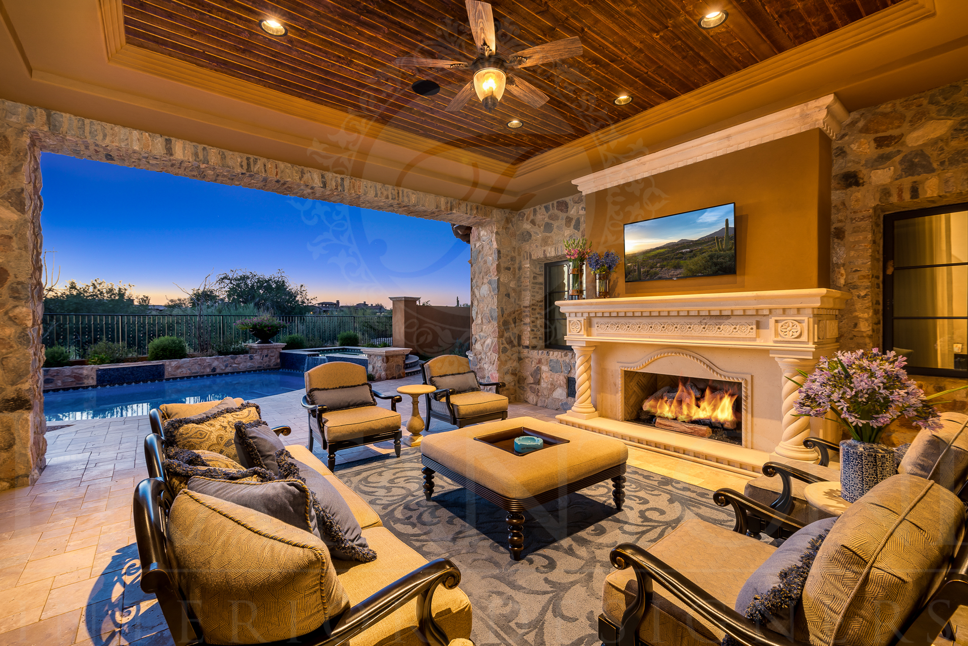 Patio with seating, fireplace, and TV overlooking a pool at dusk.