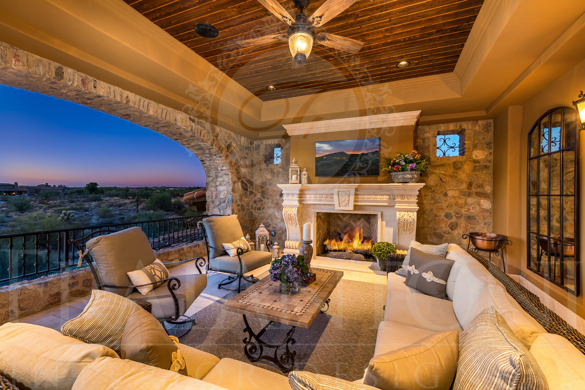 Outdoor living room with fireplace, beige sofas, stone walls, and sunset view.