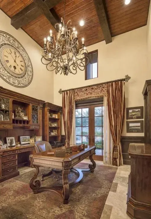 Elegant home office with ornate desk, bookshelves, large clock, and chandelier.