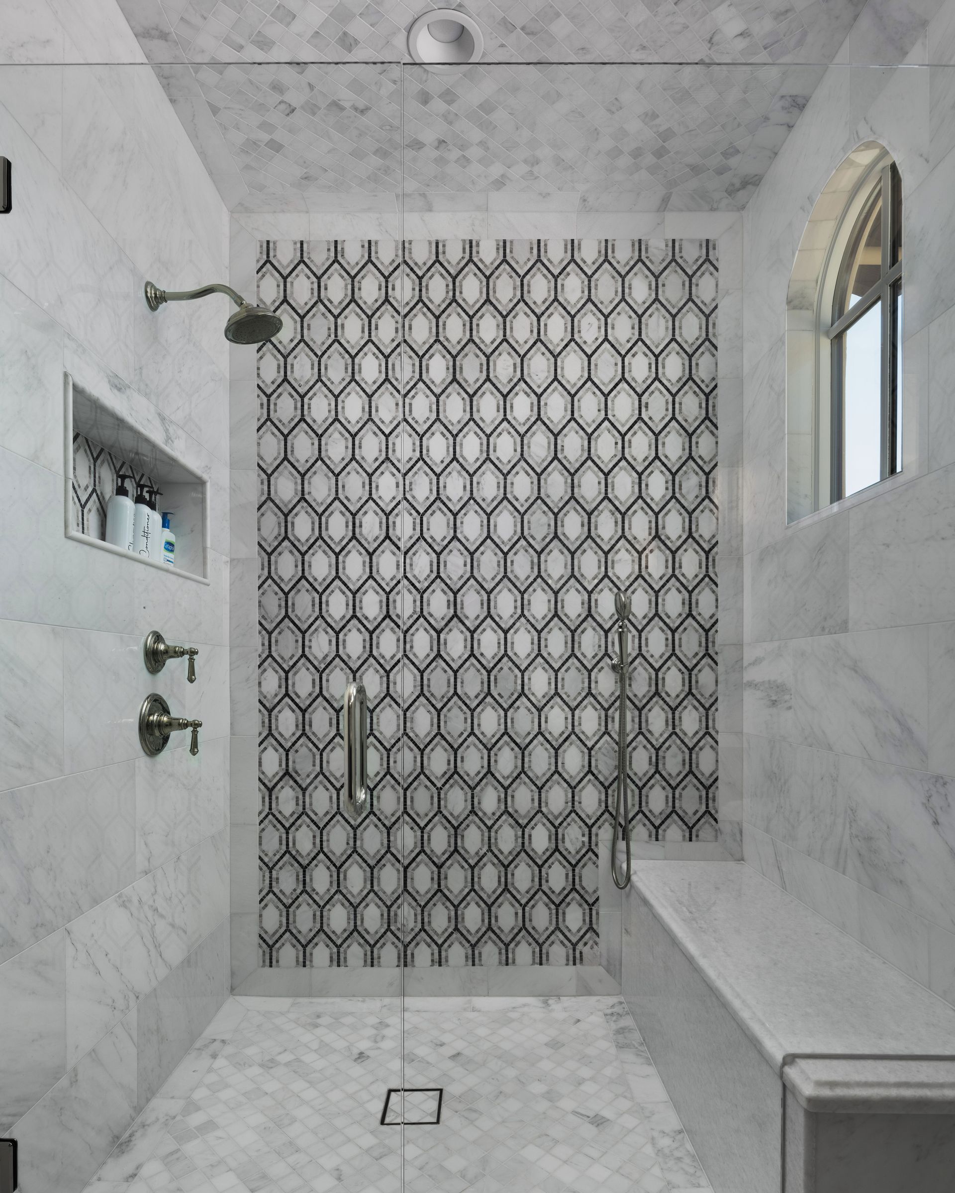 Marble-tiled shower with a geometric tile accent wall, built-in bench, and a small arched window.
