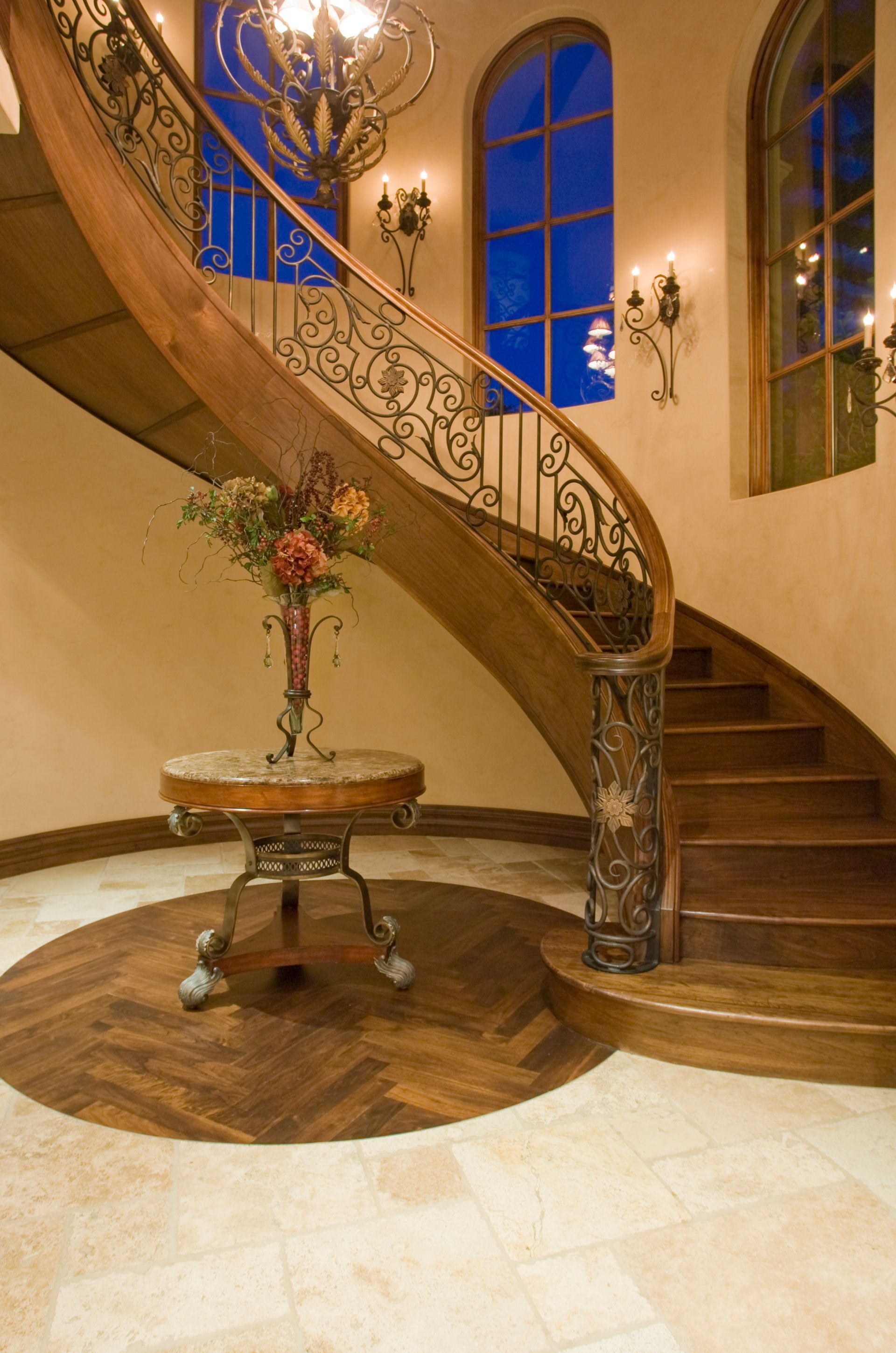 Elegant curved wooden staircase with decorative wrought iron railing, table, and arched windows.