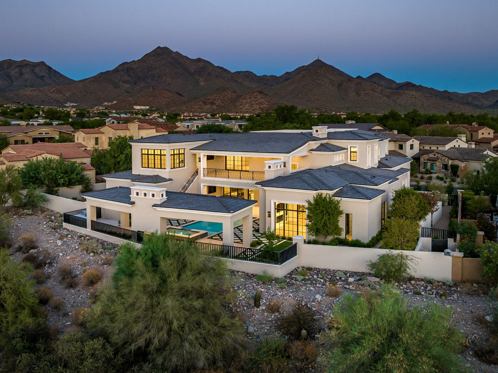 Modern mansion at dusk, beige exterior, dark roof, pool, mountain backdrop.