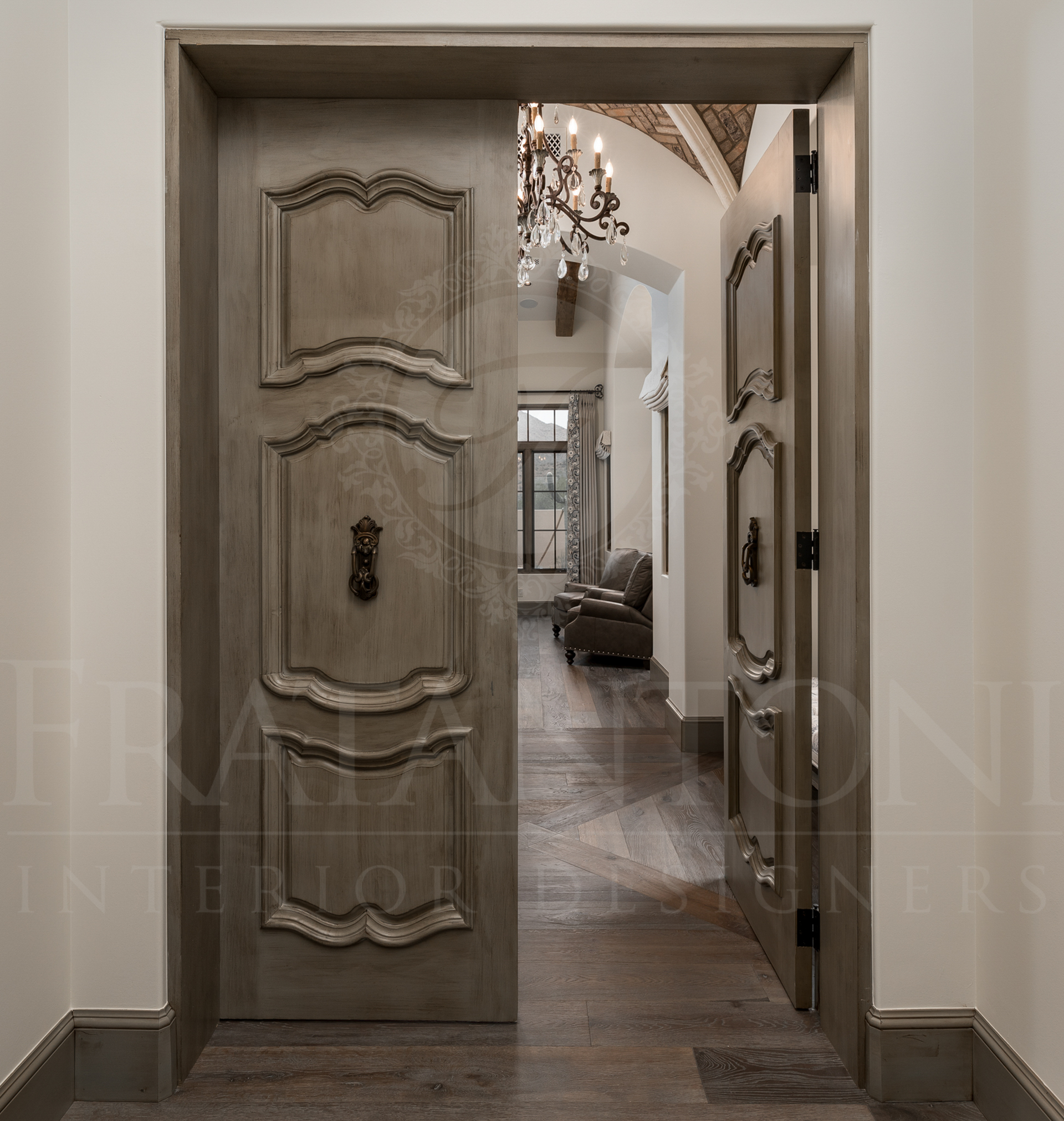 Open, ornate gray wooden doors leading to a hallway with a chandelier and bedroom furniture.