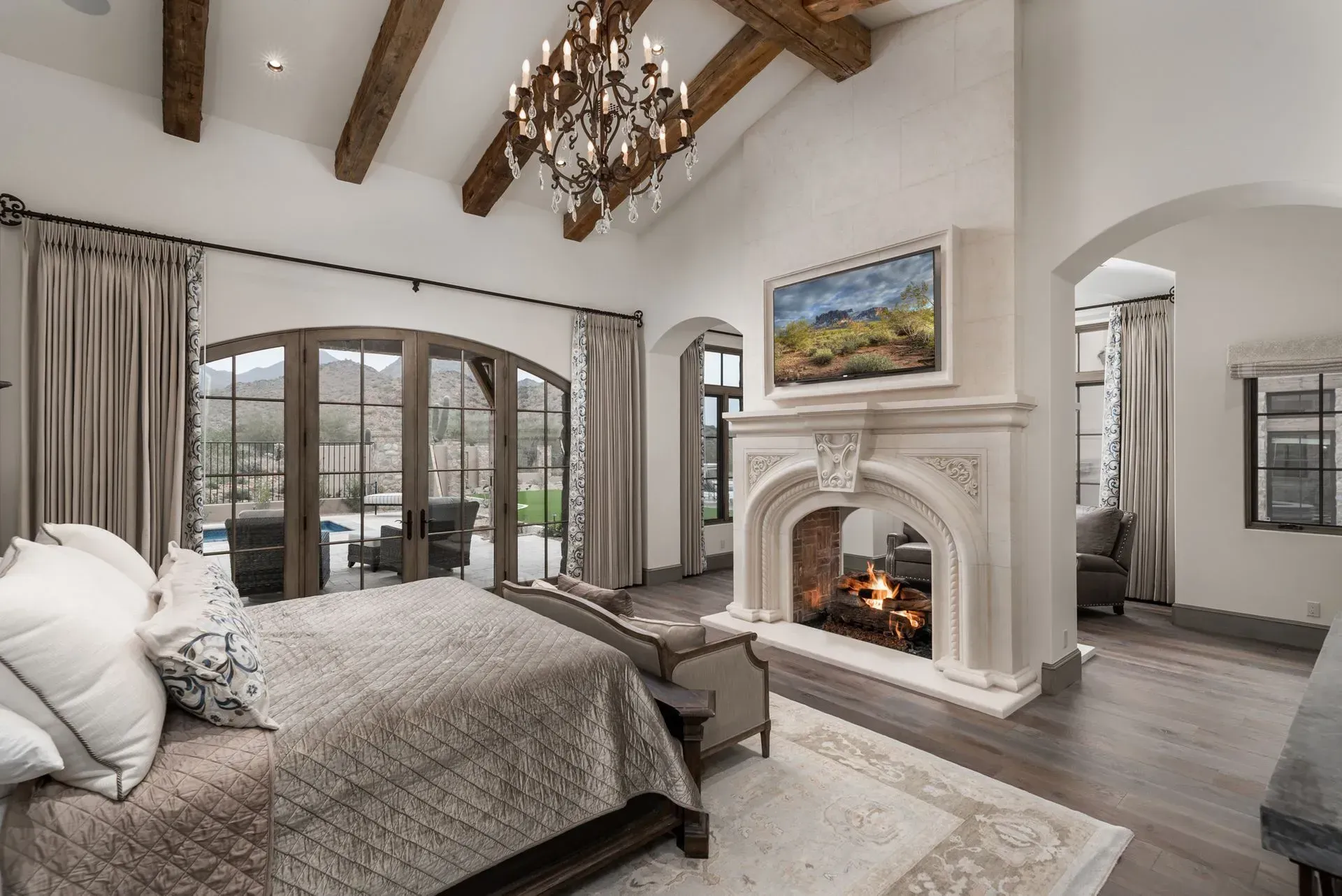 Luxurious bedroom with fireplace, arched windows, wooden beams, chandelier, and a bed with neutral linens.