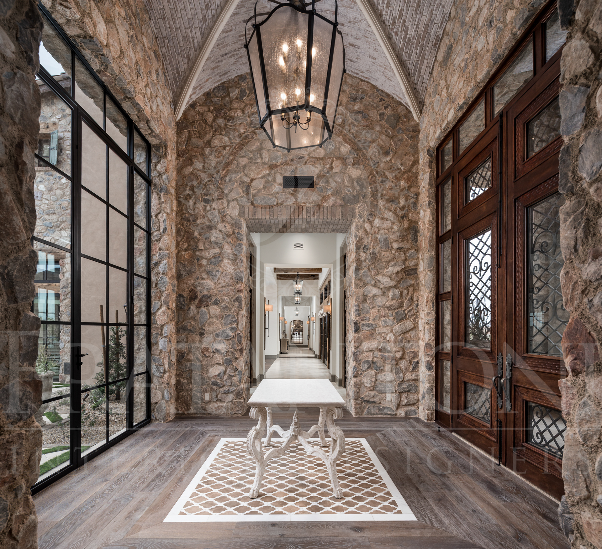Stone arched entry with large glass doors, dark wood door, ornate hanging lantern pendant, stone vaulted ceiling, and patterned floor.