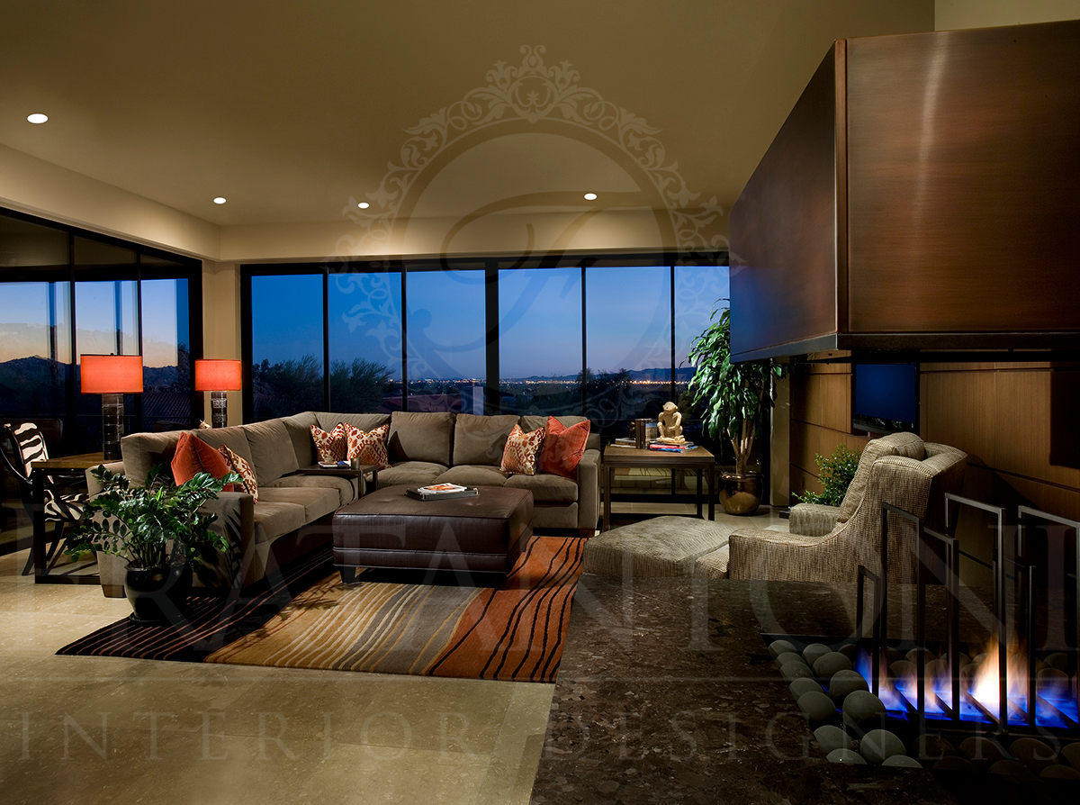 Modern living room with a large sofa, ottoman, patterned rug, and a fireplace, viewed against a dusk cityscape backdrop.