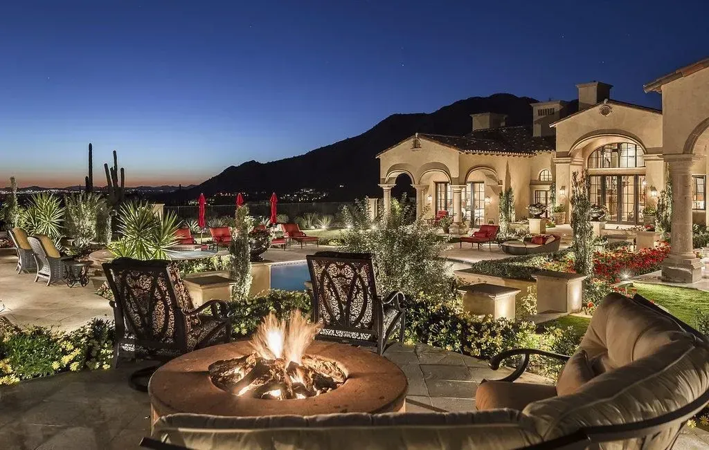 Outdoor patio with fire pit, seating, pool, and a mansion at dusk.