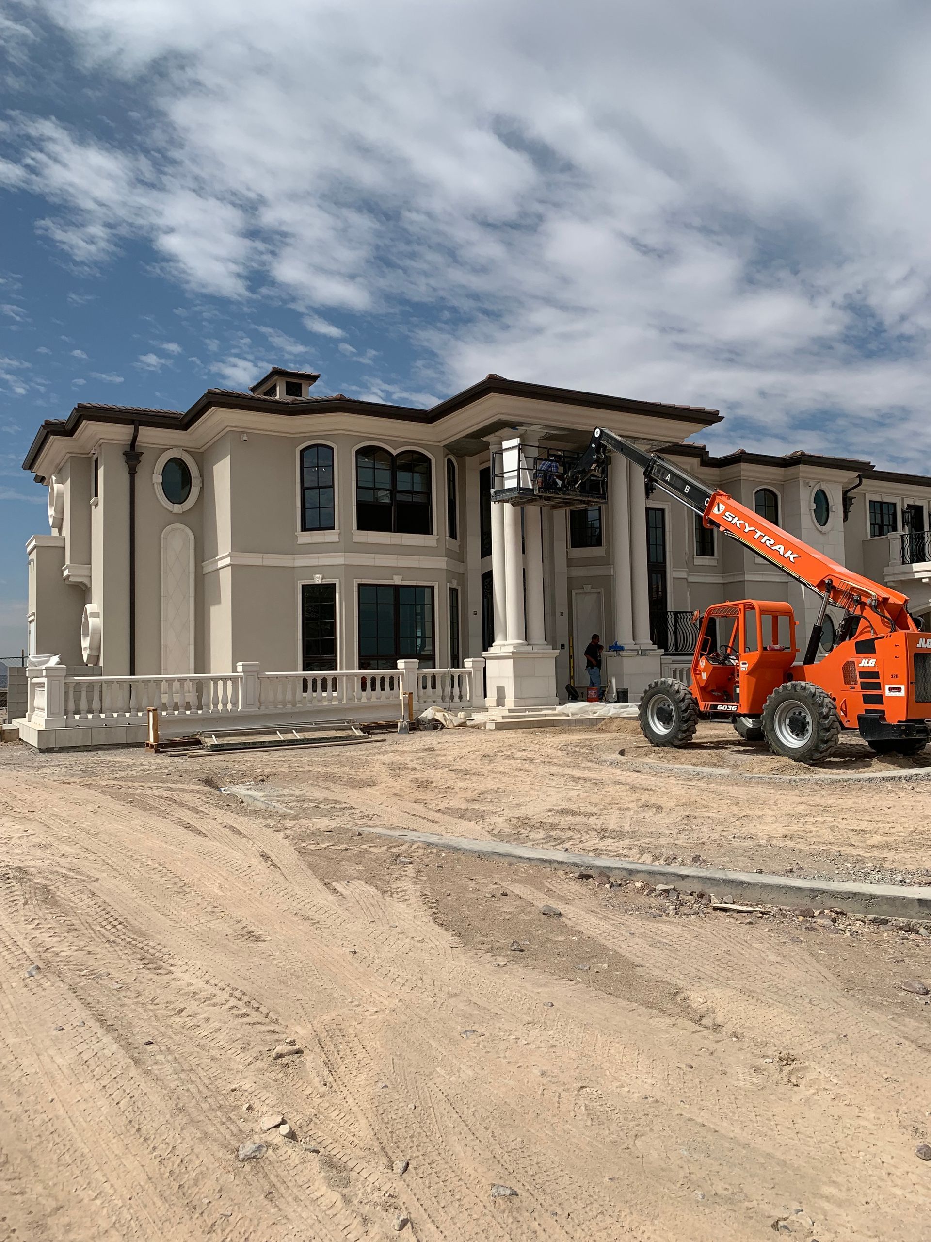 Large beige house under construction; orange lift at the entrance. Blue sky and dirt foreground.
