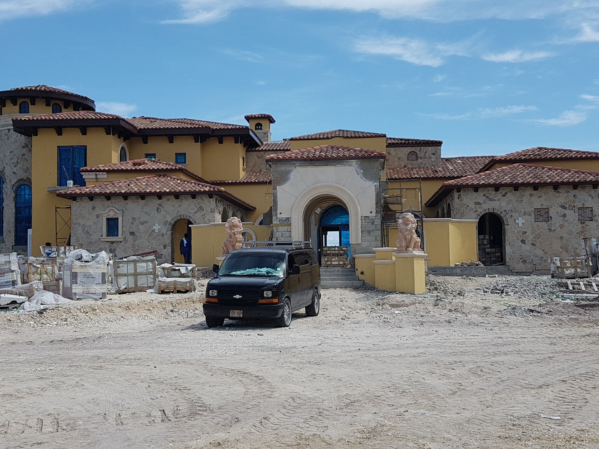 Black van parked in front of a large, unfinished stucco house with red-tiled roof under a blue sky.