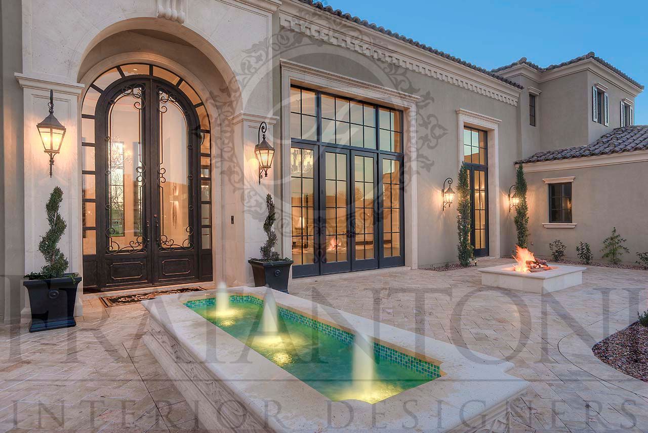 Luxury Mediterranean-style house exterior with arched iron glass doors and a small lit fountain, brick paved courtyard, fire pit, and exterior lighting at dusk