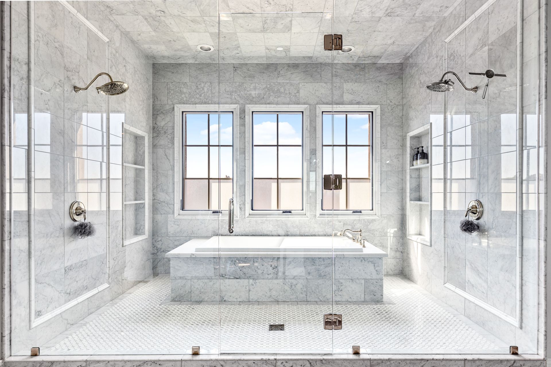 Luxurious bathroom with marble walls and floors, two glass-enclosed showers, a centered bathtub, and windows.