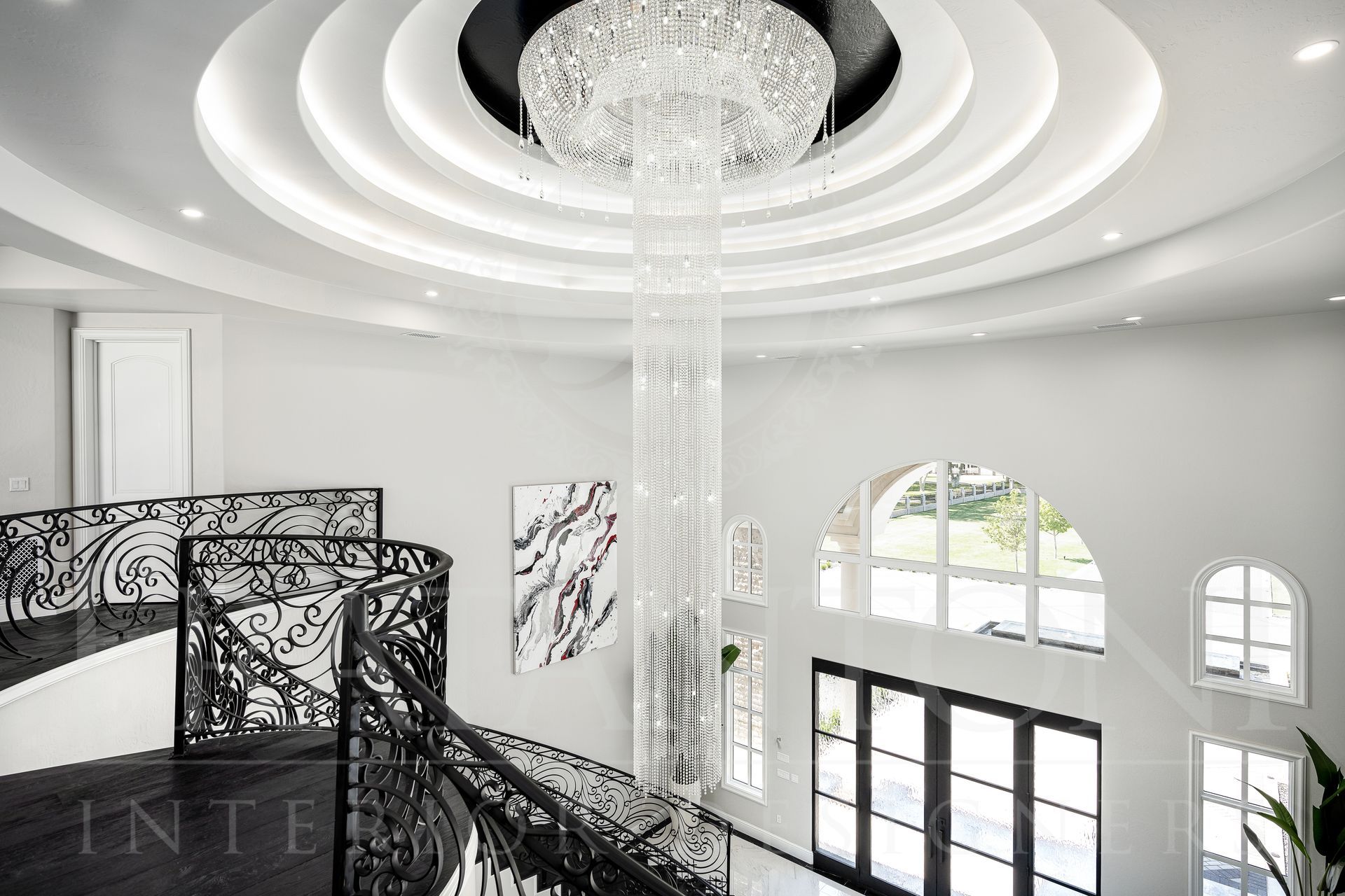 Grand, bright foyer with a large crystal chandelier, sweeping staircase, and high arched windows.