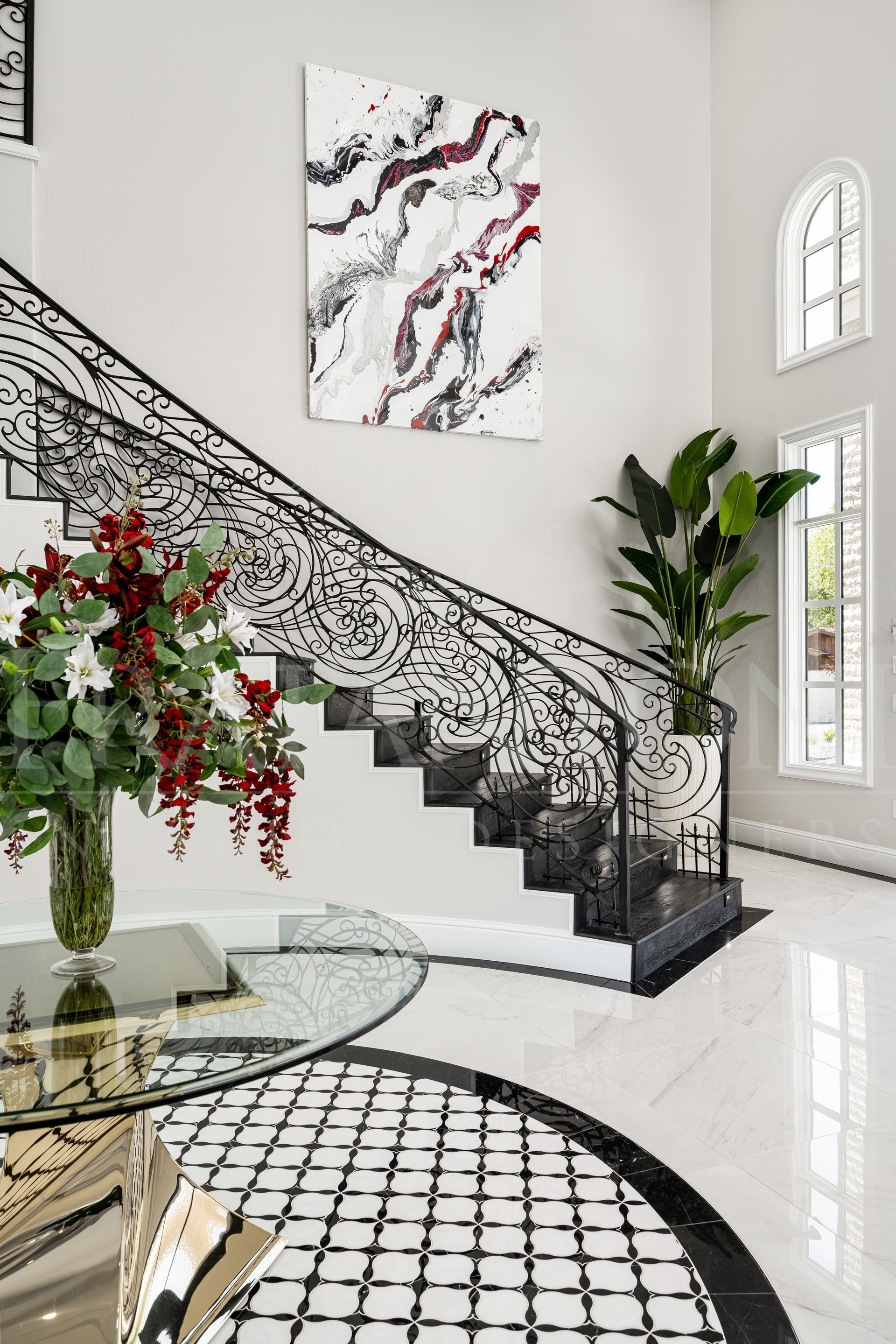 Elegant staircase with black ironwork railing, floral arrangement, abstract art, and large potted plant in a bright entryway designed by Fratantoni Interior Designers