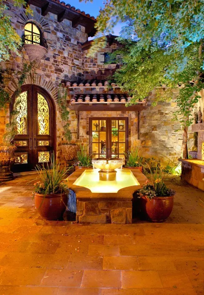 Stone courtyard at night with a fountain, potted plants, and arched doorway.