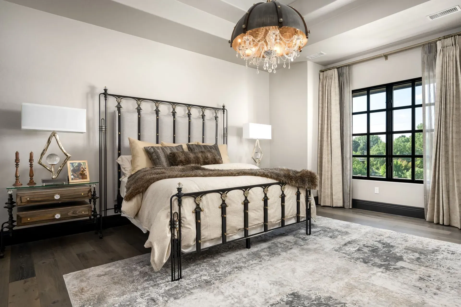 Bedroom with a black metal bed, ornate nightstands, neutral rug, and a large window with drapes.